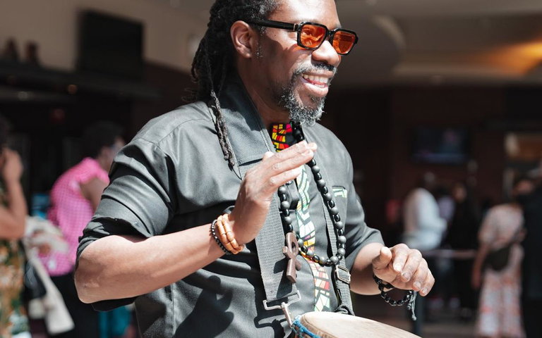 A smiling Black man wearing sunglasses is playing a djembe drum in the midst of a group of people
