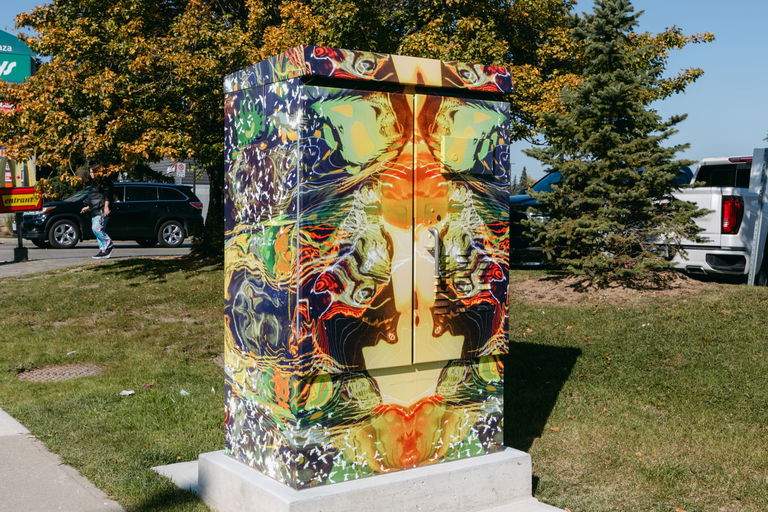 A traffic signal box wrapped in colourful vinyl. The traffic signal box is on grass, beside a cement sidewalk.