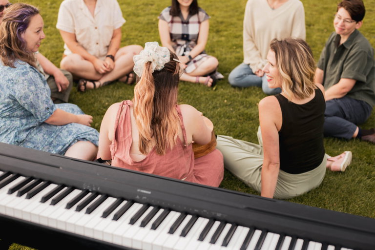 A group of people sit in a circle on the grass and smile, there is a keyboard in the background
