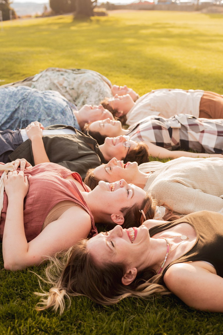 6 women and non binary people of caucasian and asian heritage and various ages lie on their backs on the grass with their eyes closed, laughing