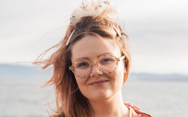 A caucasian woman wearing round gold-speckled glasses wears a pinks shirt and stands by the sea