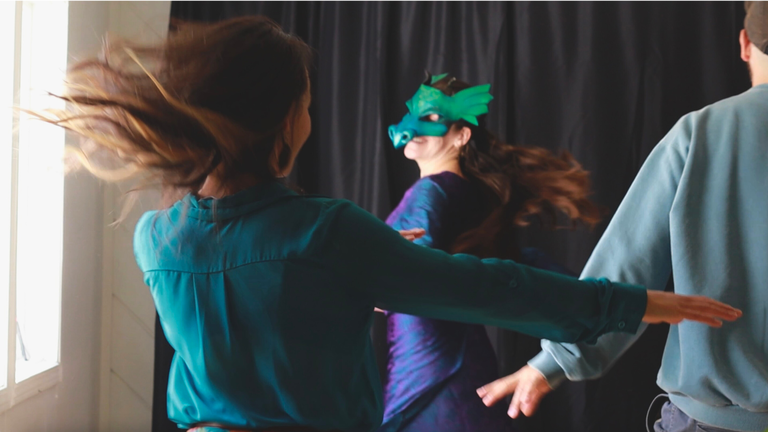 Three people dance in a circle, their hair and clothing in motion. One dancer, centered and facing the camera, wears a vibrant green dragon mask and a purple dress. The other two dancers, one in a teal shirt and one in a light blue sweater, are partially visible from behind, their arms extended outward. The background is a dark curtain, contrasting with the bright light coming from a window on the left.