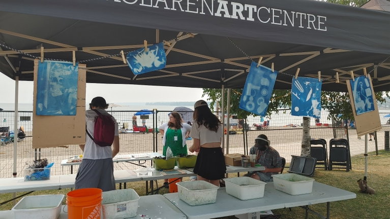A wide angle shot of several people working under an outdoor festival tent owned by the MacLaren Art Centre creating and helping to create and dry cyanotypes. They are surrounded by work tables and many water rinsing stations.