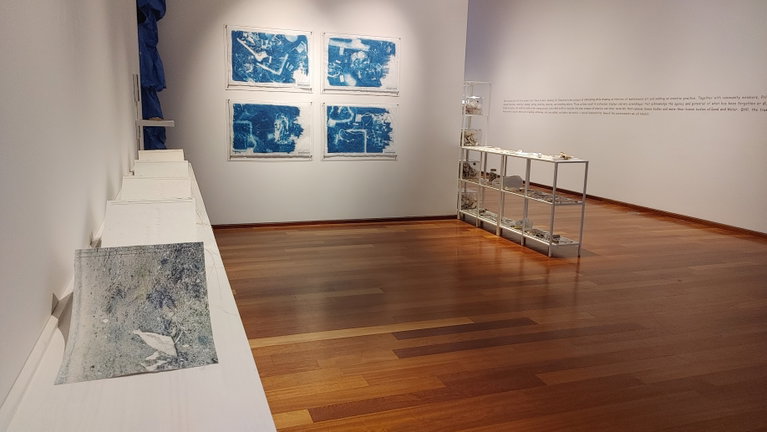 Installation shot of an exhibition by Jill Price including a long white shelf topped with a handmade accordian book work on handmade paper, four large cyanotypes on architectural drafting paper, and an arrangement of white shelves filled with white trash collected from the beach.