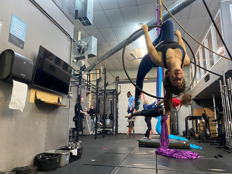 Busy gym with woman on hoop in the foreground and a woman practicing on silks behind her. Juggling and other activities are happening behind her.