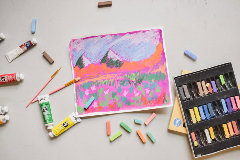 An aerial view of a table with a bright painting of mountains and flowers, with various art supplies scattered around it.