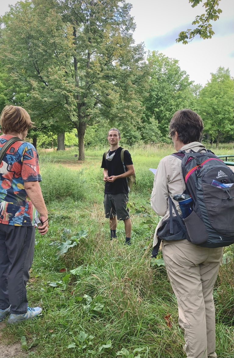 Young man standing in front of a group of people while leading a nature walk in a park.