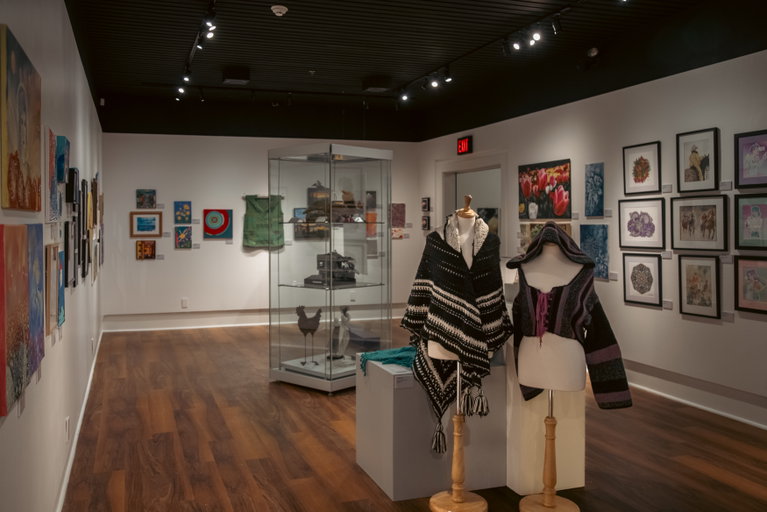 Art gallery featuring framed artwork, textile pieces, and a central display case in a modern exhibition space with wood floors.