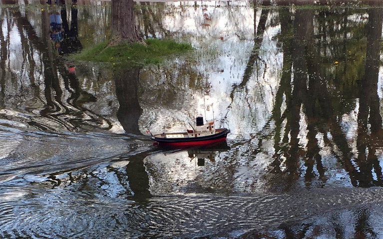 A toy boat in water of a flooded park. In the reflection of the water, an adult figure and child are seen with a remote control.
