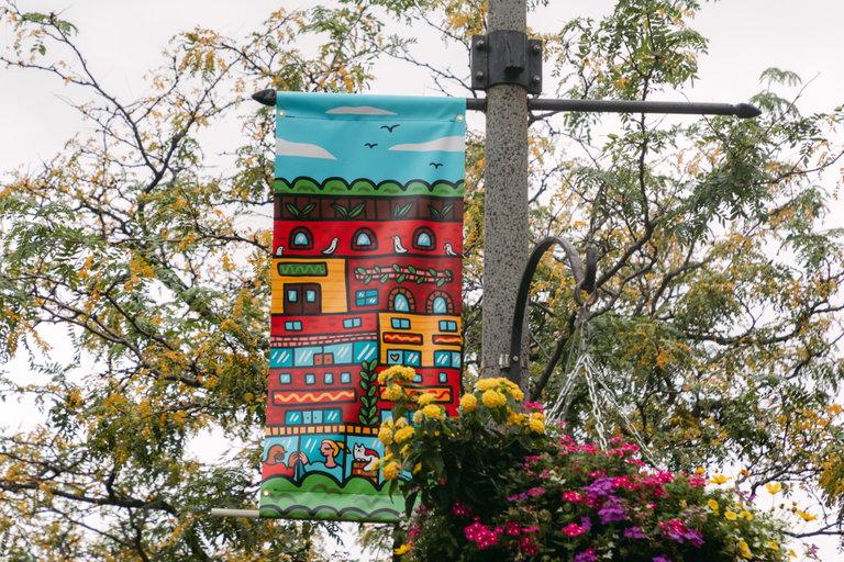 An pole banner hanging up streetside. The artwork on the pole banner is of red, yellow, and blue houses and shops.