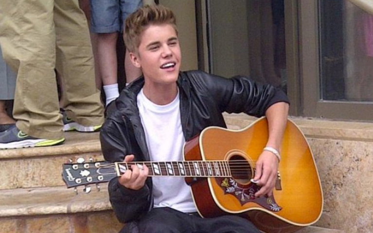 Justin Bieber sits on the steps of the Avon Theatre and performs a song on his guitar.