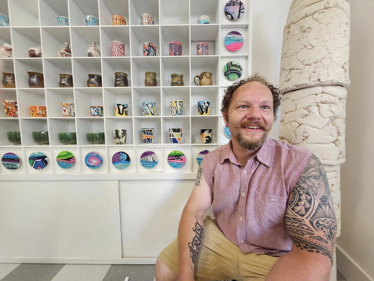 Métis artist Denny Provost sits in from of wall showcasing ceramic cups