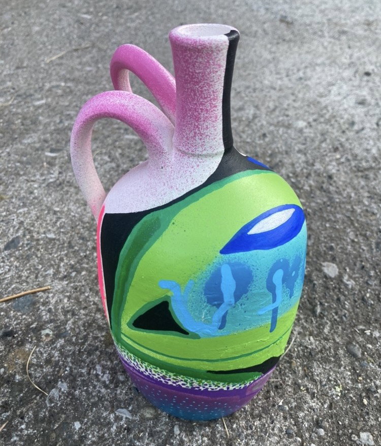 A ceramic jug with wide handle and thin spout covered in spray paint, drips and solid colors of black, green and blue