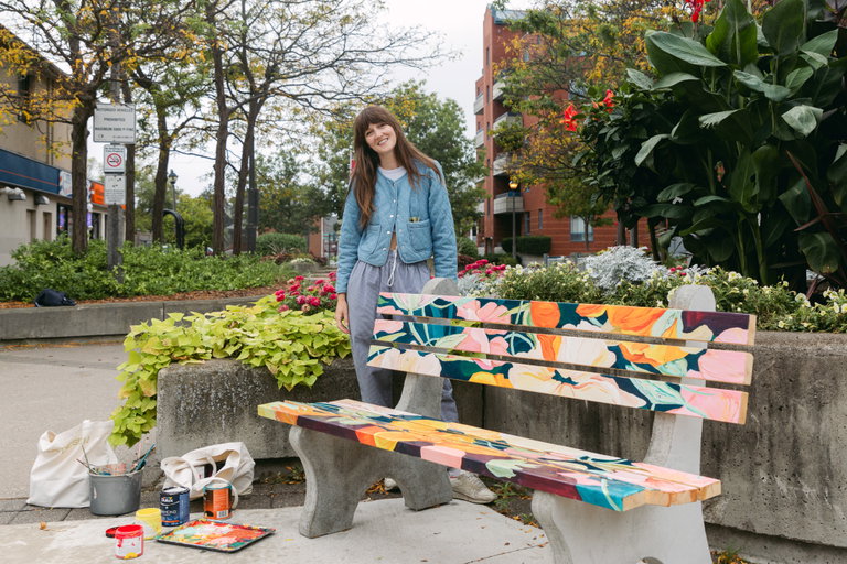 Artist Kayla Whitney standing beside a bench with wooden slats that she hand-painted with ornate flowers. Paint supplies are on the ground below the bench. including paint cans and a palette.