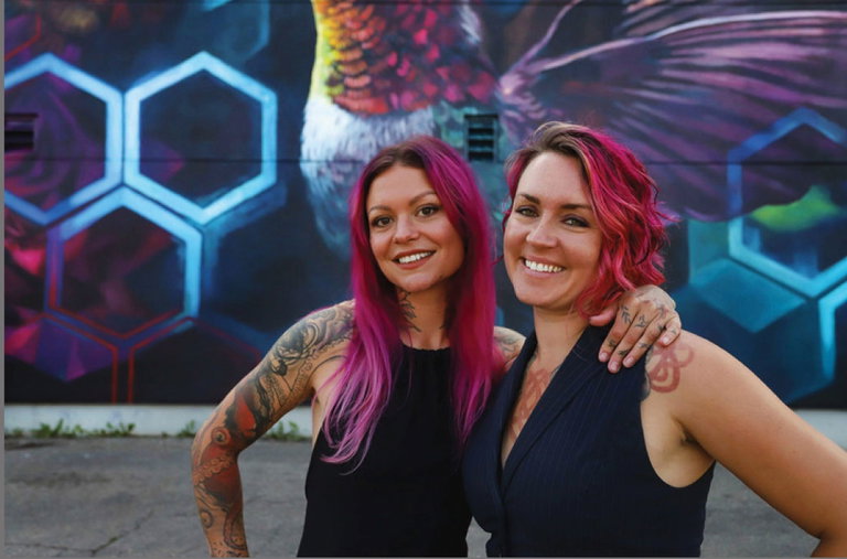 LALA Art - Lacey Jane Wilburn and Layla Folkmann pose in front of a large, colourful mural with a honeycomb design