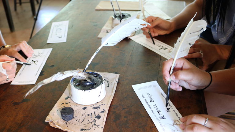 Close up view of three people practicing their penmanship using feather quill pens and ink.