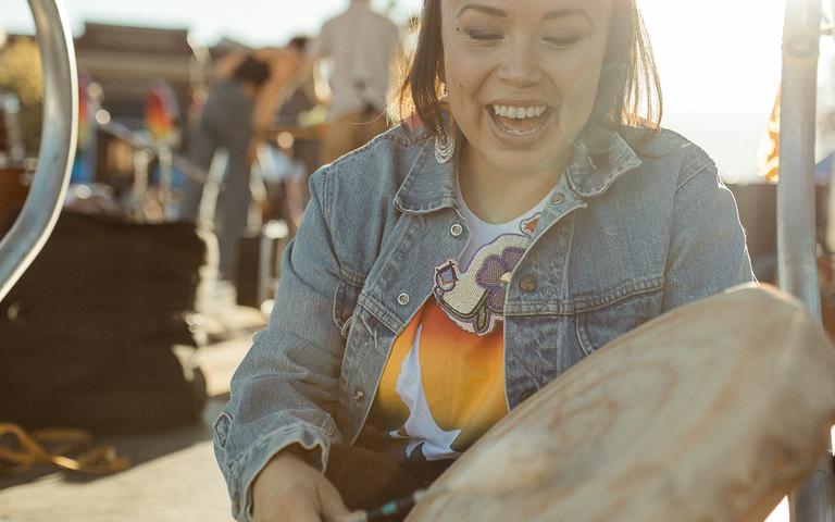 A non-binary person sits on the steps of a stage. They are wearing a jean jacket over a white t-shirt. They have beaded rainbow earrings and Indigenous beadwork. They are playing a drum as the sun shines on them from behind them. In the background technicians are setting up a piano.