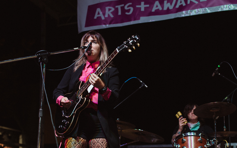 A feminine presenting person with shoulder length blonde hair plays guitar while singing into a microphone. She is on stage, wearing a pink button up, a black blazer, black shorts, and fishnet stockings. Behind her, a brunette drummer his oldering drumsticks while sitting at a drum set. Behind and above is a banner that reads "Salmon Arm Pride Project Arts + Awareness Festival"