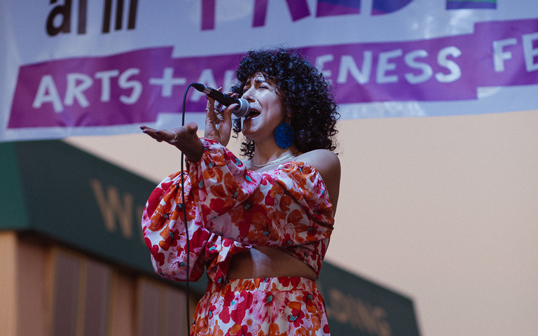 A femme presenting person sings into a microphone on stage. She has dark curly hair and is wearing a two piece orange, pink, and white flower print dress. She wears a silver ring on her left forefinger and large round blue earrings. Behind her is a banner that reads "Salmon Arm Pride Project Arts + Awareness Festival"