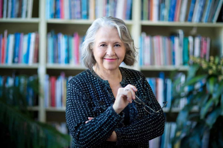 a woman shoulder-length, slightly wavy silver hair, smiling - she is standing infront of shelves of multicolored books