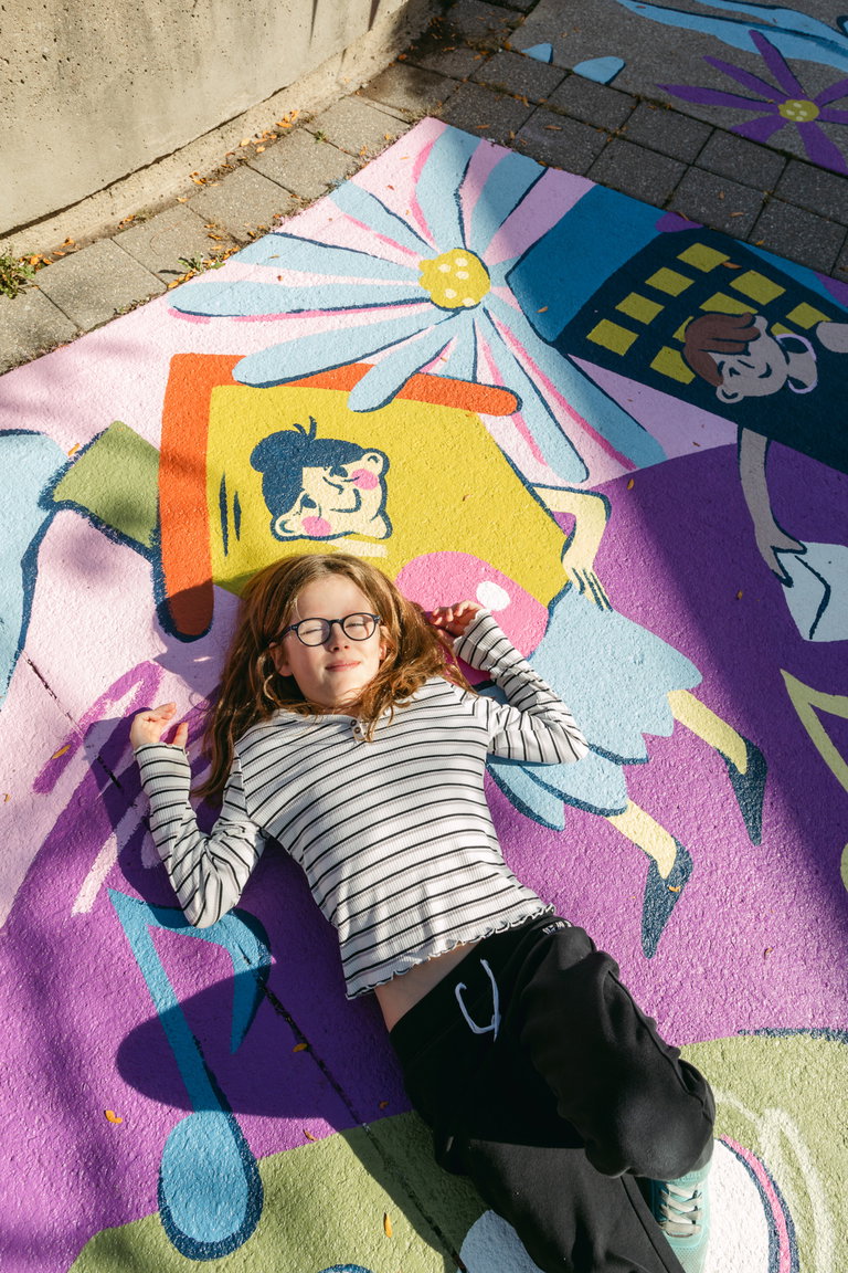 A child with long blonde hair, black-rimmed glasses, and a black-and-white striped long sleeve shirt laying on top of the painted cement mural.
