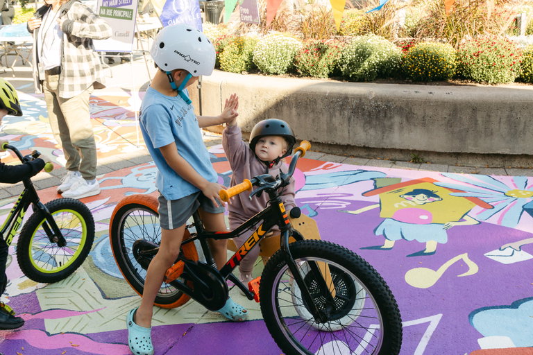 Two children on bicycles high-fiving. The children are standing on the ground, below them is a colorful mural painted on the concrete.