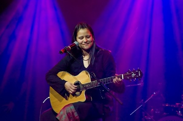 Pam sits, playing guitar, in front of a microphone, with purple lights behind her