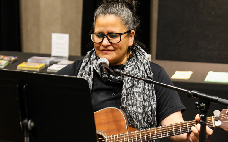 Pam sits, playing guitar, in front of a microphone