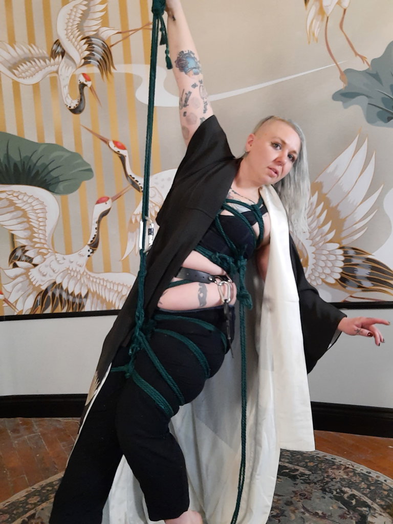 A blonde tattooed person wearing black pants and a vintage black kimono is suspended in a standing position using dark green rope. There is a mural depicting flying cranes behind them.
