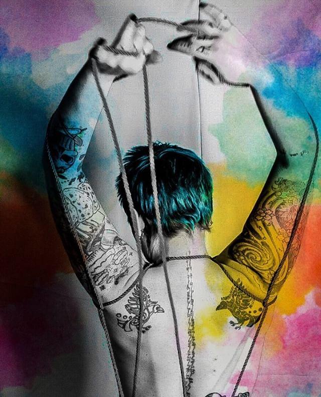 The artist faces away from the camera, in the midst of tying themselves with a piece of rope. The main image is in black and white with splashes of rainbow colour framing the exterior.