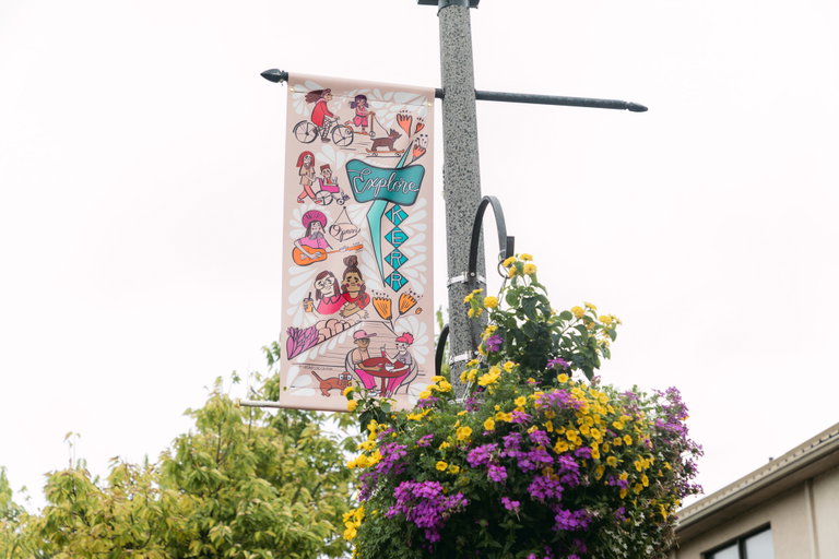 A street pole banner hanging from a metal rod. The pole banner is pink, blue, and orange. It illustrates women and children drinking coffee, going for a walk, and riding bikes down Kerr Street.