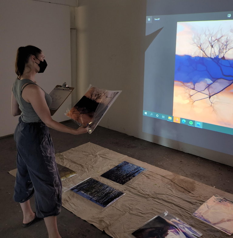 A woman wearing standing in front of a work of art holding a photograph and a clipboard.