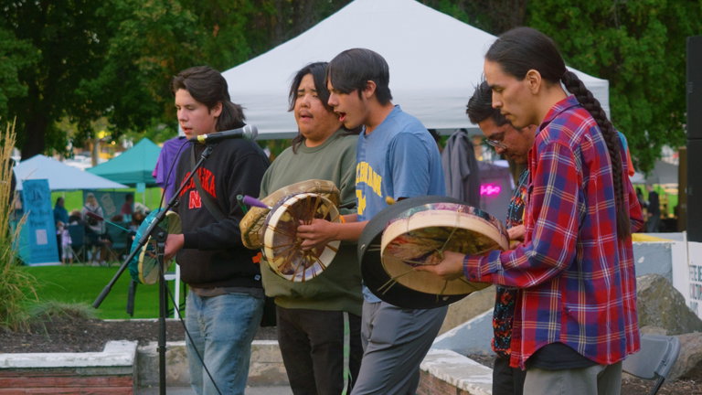 A group of six Indigenous men perform at an outdoor event, singing and playing hand drums. They stand in a line near a microphone, deeply engaged in the music. Behind them are white tents and canopies, with people gathered in a grassy park setting, suggesting a community or cultural festival.