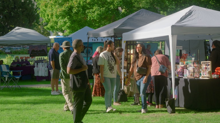 People browse artisan vendor booths under white and grey canopy tents at an outdoor festival on a sunny day. The booths display a variety of handmade items, including jewelry and crafts. Attendees chat and walk on the grassy park grounds, surrounded by trees and dappled sunlight.