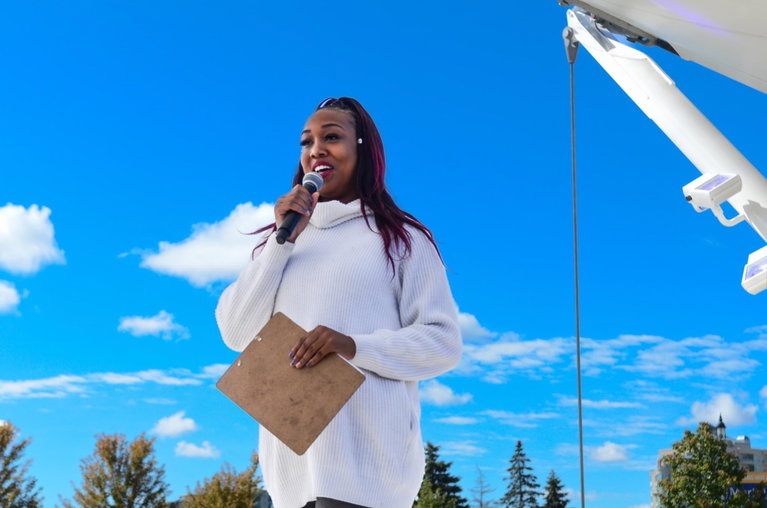 The Poet Laureate is shown on stage, holding a mic, smiling, against a bright blue sky.