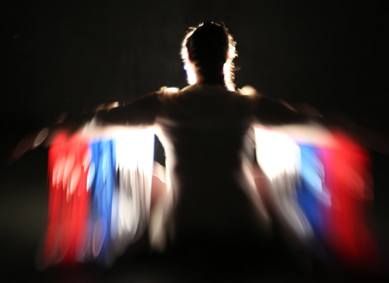 A blurry video-still shows a feminine figure with intense back-lighting- turning the figure into an outline or silouette against a black background. Coloured fringe hangs from the figure's outstretched arms, in red, blue, and white. The figure appears to be in motion.