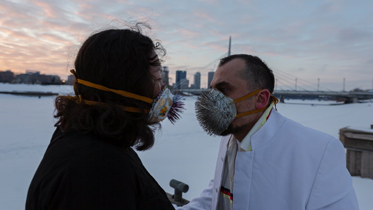 Two people face each other, one male presenting, the other female presenting. The woman has long, black hair and is wearing black. The man has short brown hair and is wearing a white suit jacket. They both wear medical masks that have been decorated with cloth and stitching. Sticking out of the masks and pointing at the other person are many porcupine quills. They stand outdoors with a sunset and city-scape in the background. They are surrounded by snow.
