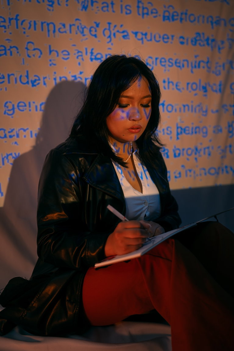 A filipino artist sitting on the ground writing in a note book. The artist is in front of a backdrop with rows blue text.