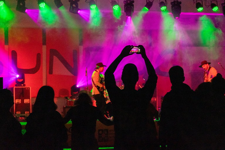 Musicians perform on a brightly lit stage with green and purple spotlights, while silhouetted audience members watch and one person records the show on a smartphone.