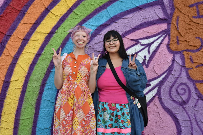 Two ladies dressed colourfully smile and make peace signs with their hands in front of a rainbow mural
