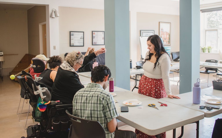 A group of people sit around a table, beading, while an instructor stands in front of them.
