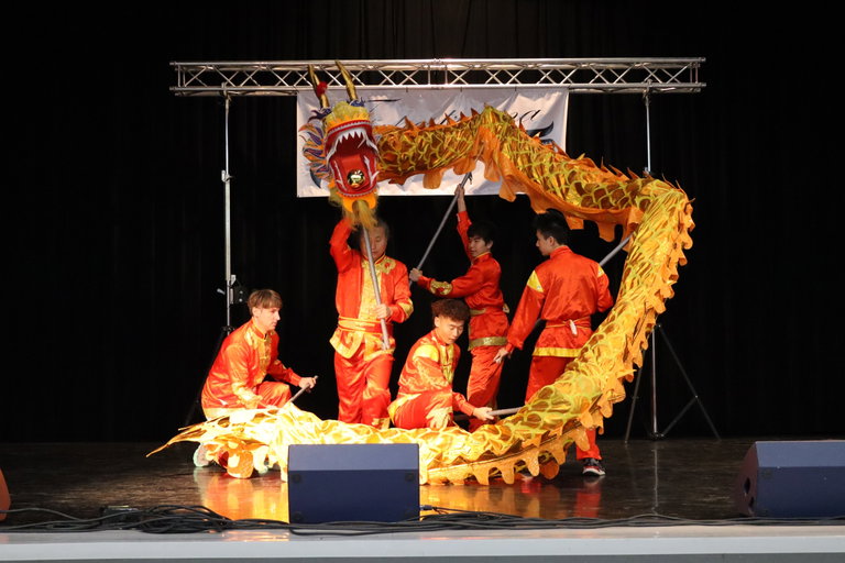 A group of five men, dance in traditional Chinese dress, red and gold in colour. They form a circle, each holding up a part of a yellow and gold dragon made out of fabric.