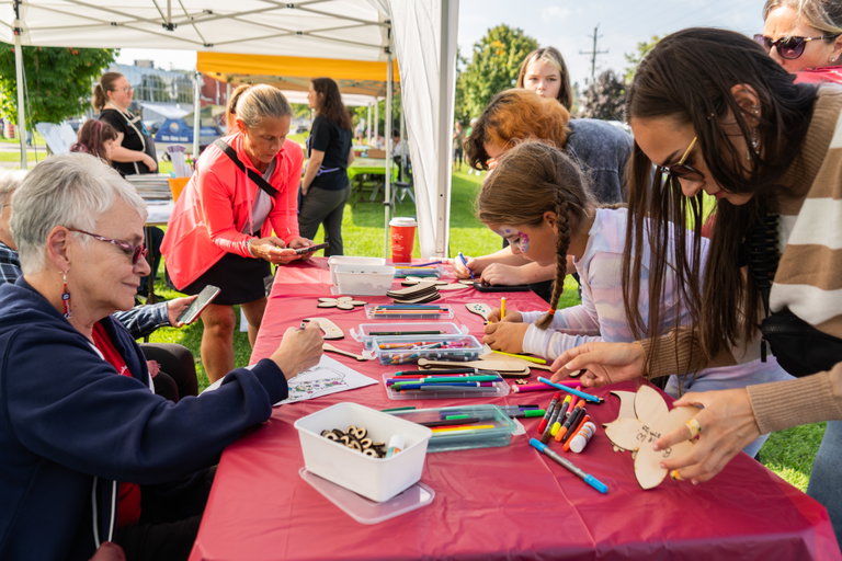 A group of people gathered under a white outdoor tent at a community event, sitting around a red-covered table and decorating small wooden crafts with colorful markers. Containers of markers and supplies are spread across the table, while additional attendees and tents are visible in the grassy background on a sunny day.
