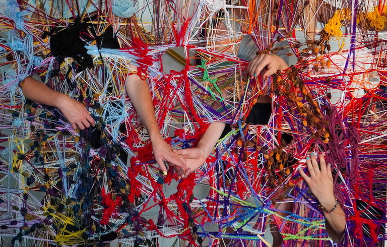 A group of multiple hands intertwine with a wall full of colourful yarn weavings.