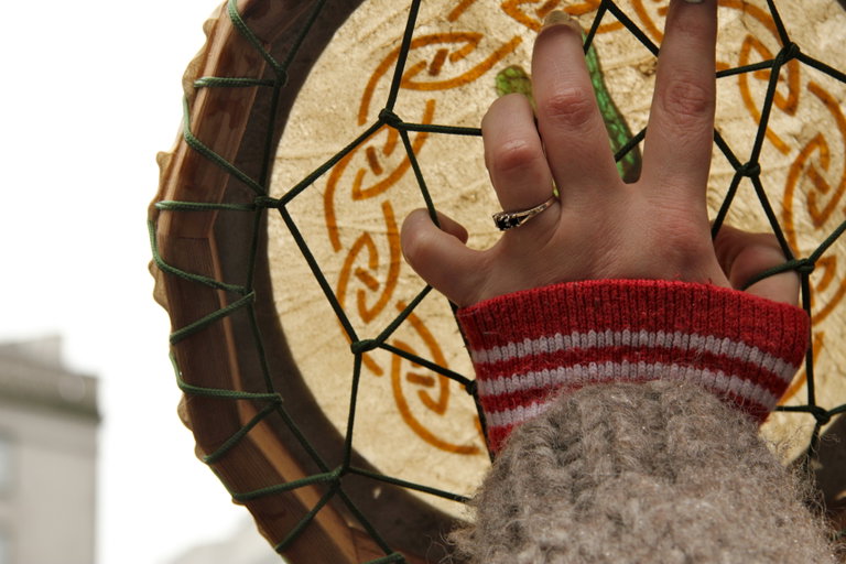 A hand with a ring on the fourth finger, coming out of a red and white striped sleeve and beige wool sweater, holding an Indigenous drum with fingers gripping a web of strings incasing the drum.