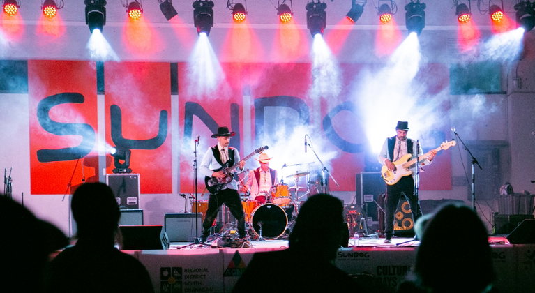 The Pseudos performs energetically on stage at the Sundog Festival of Arts and Culture. In the background, a large "SUNDOG" banner hangs across the stage, complemented by theatrical smoke effects. A crowd of people is gathered in front of the stage, enjoying the live music performance.