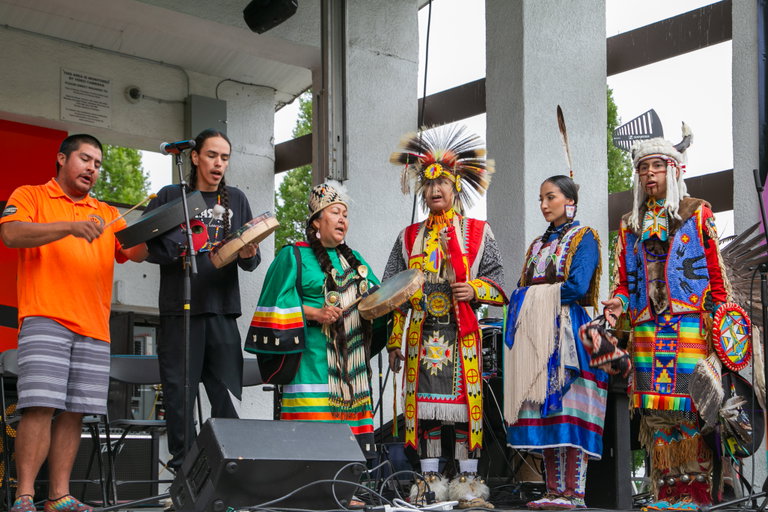 Four Syilx Okanagan singers and drummers stand together, wearing vibrant traditional regalia. Their attire includes intricately beaded garments, feathered headdresses, and handcrafted accessories. They hold traditional drums and sing, showcasing their cultural heritage and musical traditions.
